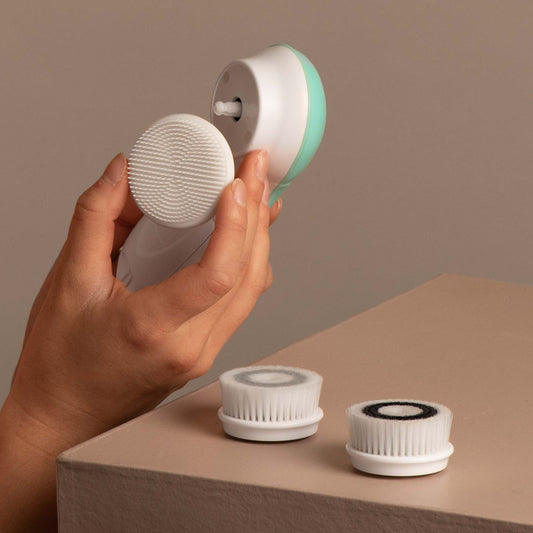 A hand attaching a silicone facial brush head to a Cora 3 device, with two replacement bristle heads on a table.
