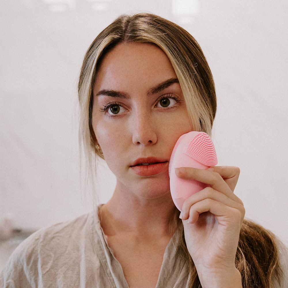 Woman using the Isla pink sonic facial cleansing brush with silicone bristles to exfoliate and massage her cheek.