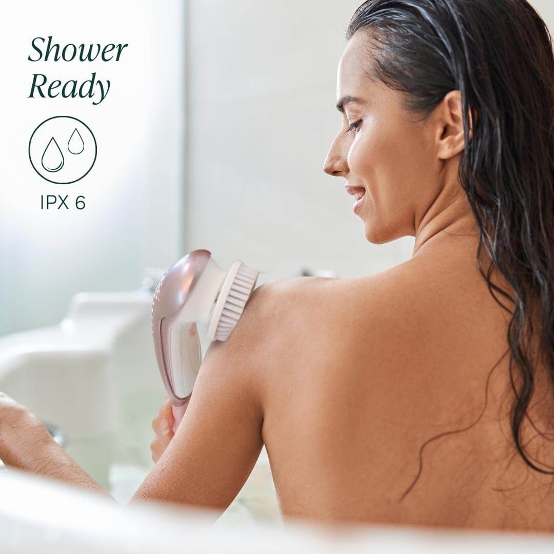 Woman in shower using Cora 7 waterproof electric body cleansing brush on shoulder. IPX6 rated for bath use.