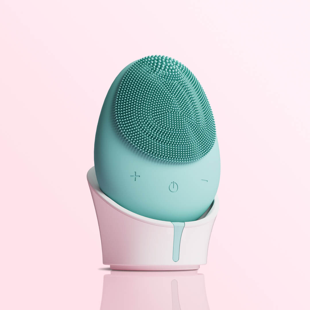 Isla mint green sonic silicone facial cleansing brush and exfoliating scrubber in a white stand against a pink background.