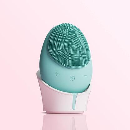 Isla mint green sonic silicone facial cleansing brush and exfoliating scrubber in a white stand against a pink background.