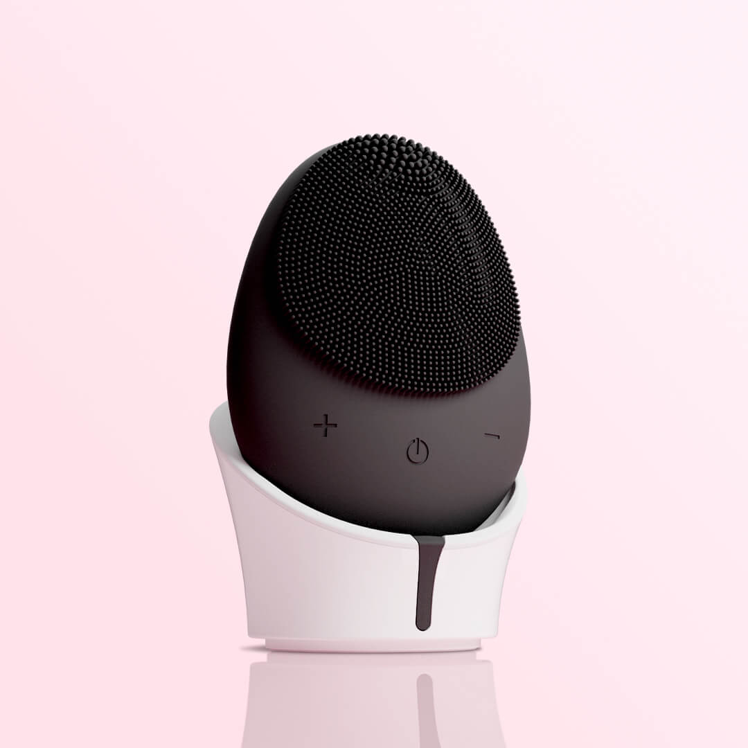 Isla black sonic facial cleansing brush with silicone bristles on a white charging stand against a pink background.