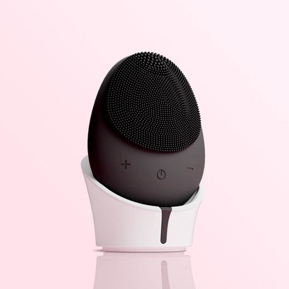 Isla black sonic facial cleansing brush with silicone bristles on a white charging stand against a pink background.