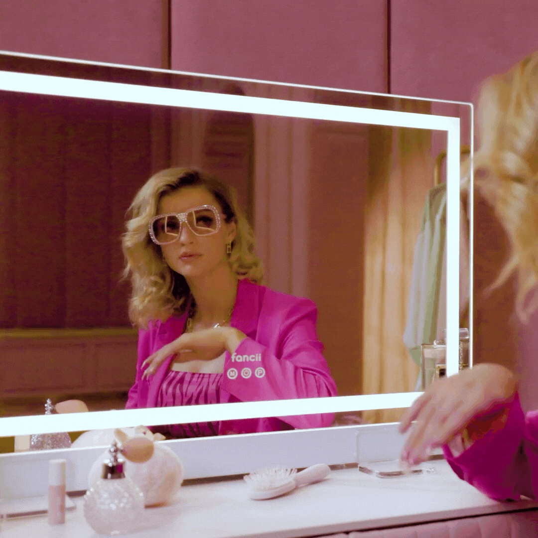 Monroe Hollywood vanity mirror with Omni-Glow LED lights reflecting a woman in pink against a glam makeup station.