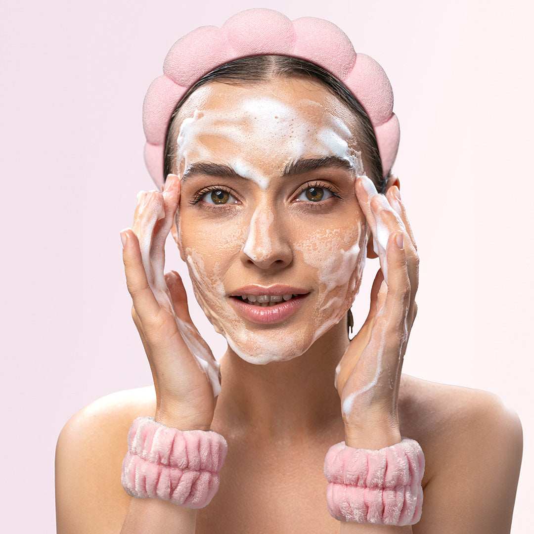 A woman washing her face with foam, wearing the pink Avery bubble spa headband and matching microfiber wristbands.
