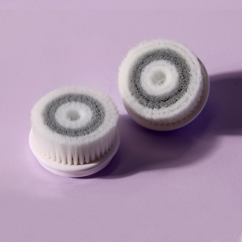 2-pack of Cora facial cleansing replacement brush heads with dual-tone grey and white bristles on a purple background.