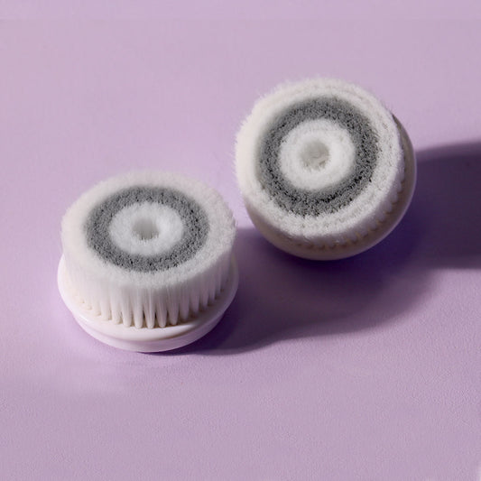2-pack of Cora facial cleansing replacement brush heads with dual-tone grey and white bristles on a purple background.