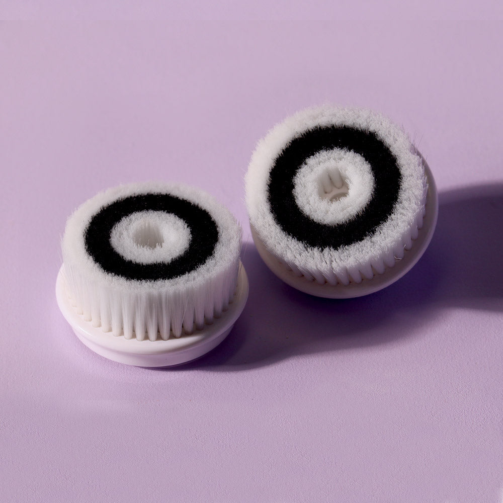 Cora 2-pack exfoliating facial cleansing brush head replacements with white and black bristles on a purple background.