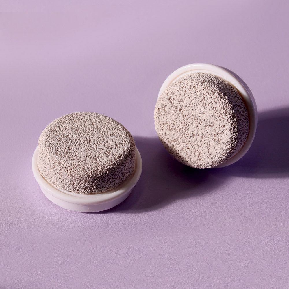 Two-pack of Cora Pumice Stone Replacement Brush Heads for facial cleansing and foot smoothing on a purple background.