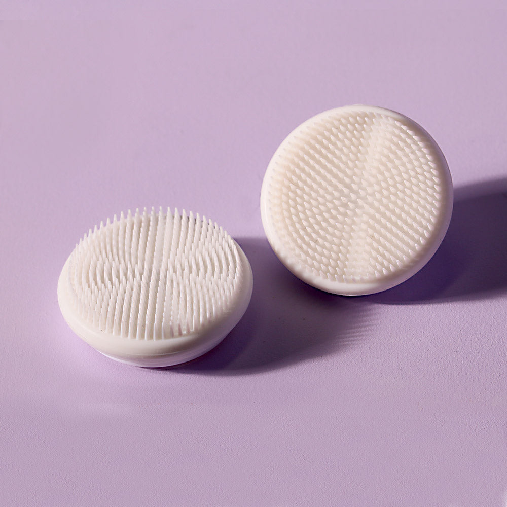 Two white silicone replacement facial cleansing brush heads for Cora devices on a purple background.